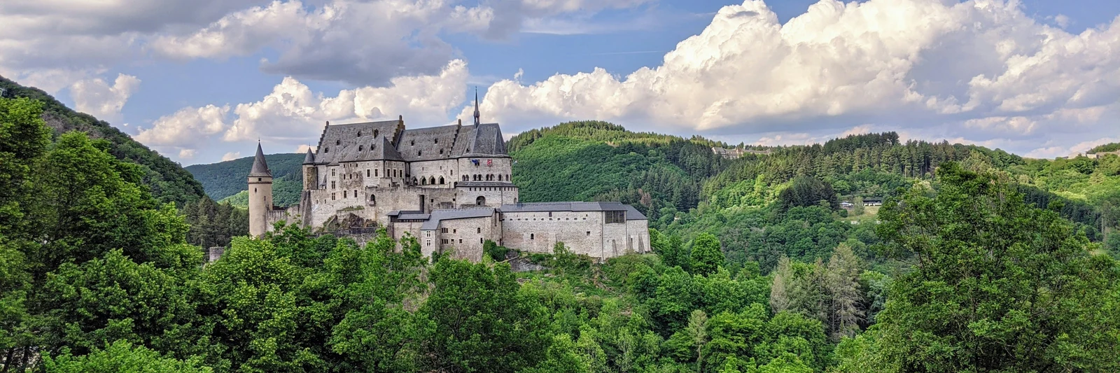 Vianden Castle surrounded by green forested hills beneath sunlit clouds, Luxembourg