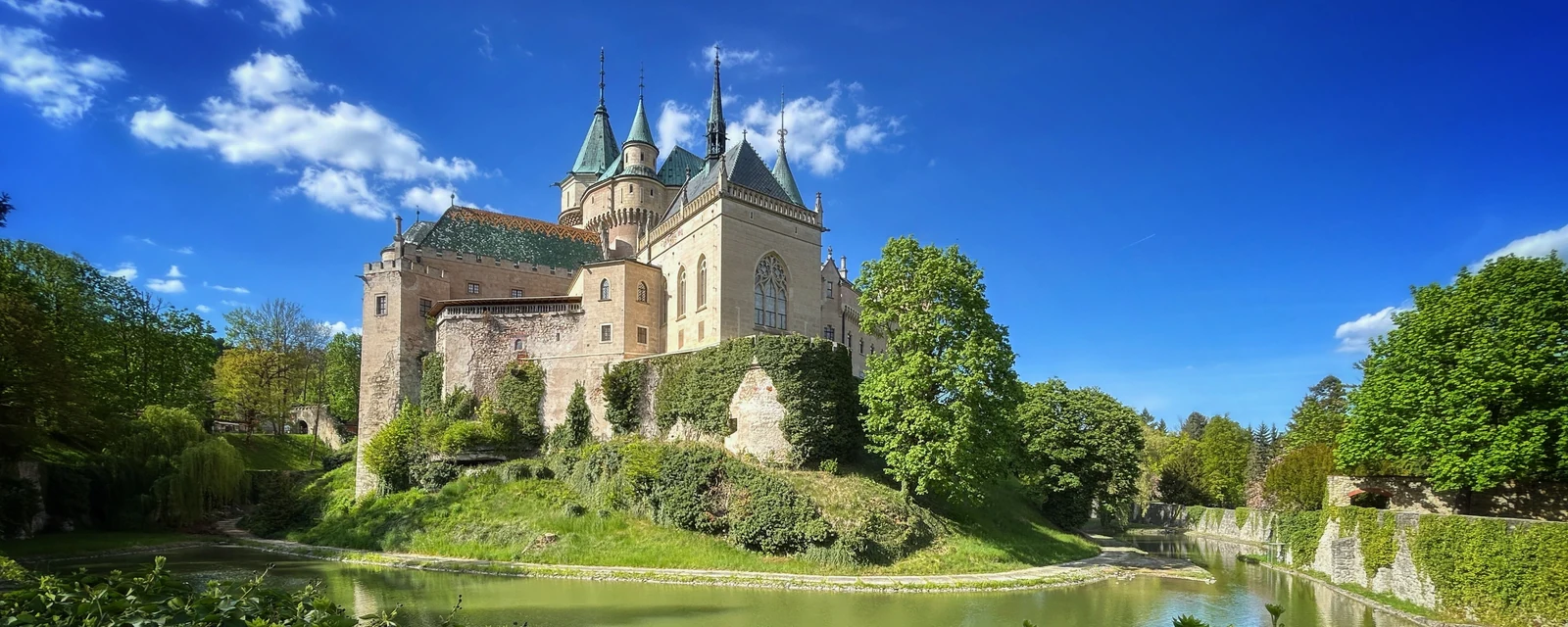 Sunlit Bojnice Castle with green gardens and a curving moat beneath blue sky, Slovakia