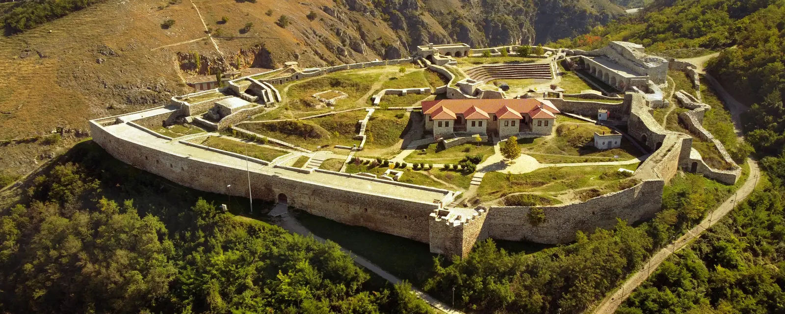 Sunlit stone ramparts and red-roofed buildings of Prizren Fortress above green mountain slopes in Kosovo